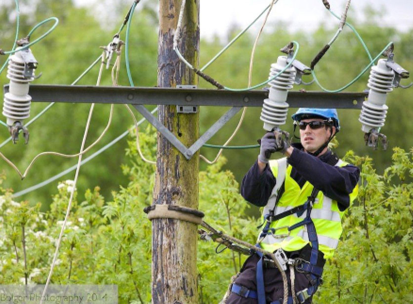 electrician with yellow hi-wis jacket and blue hard hat climbing a pole and fixing a transformer