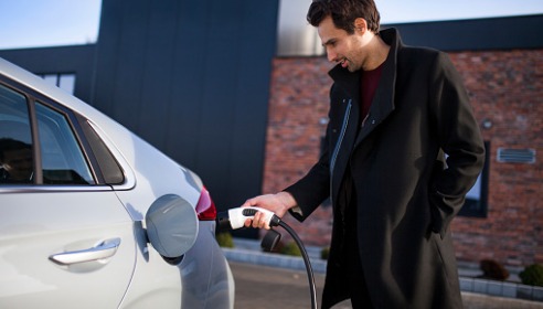 White man with brown hair and wearing a long coat holding an electric vehicle charging plug and putting it into a grey car outside a building